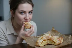 Vrouw eet vers gebakken broodje aan tafel, gebak op bruin papier, binnen omgeving.